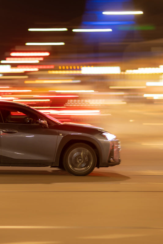 A grey car is driving at night, with streaks of red, white, and blue lights blurring past it, indicating motion.