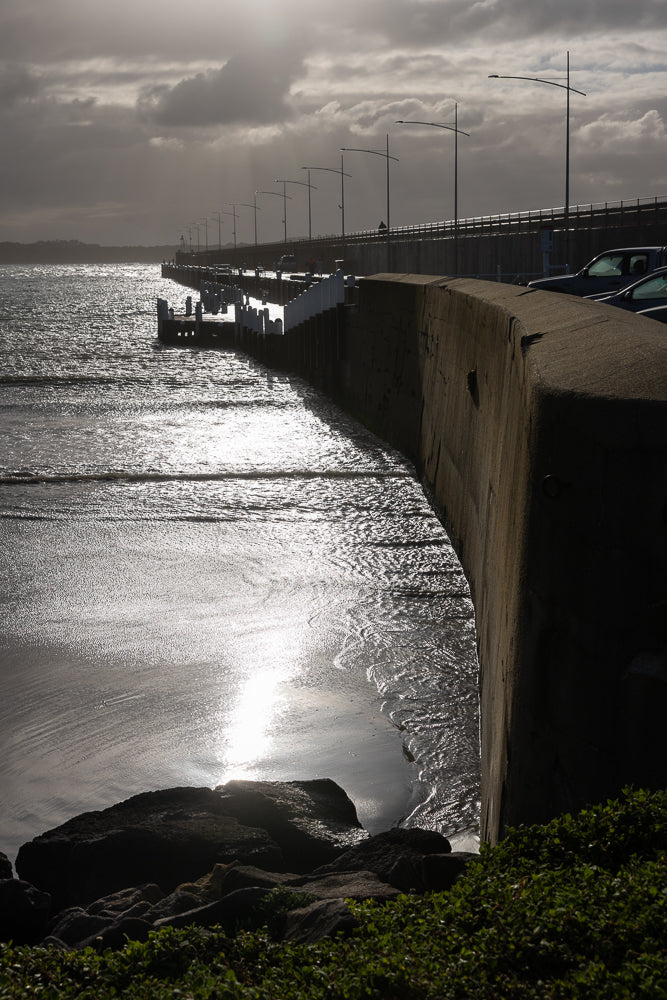A concrete sea wall curves into the distance along a beach. The sun reflects brightly off the choppy water. A pier with white posts extends into the sea. A bridge with streetlights is visible in the background.