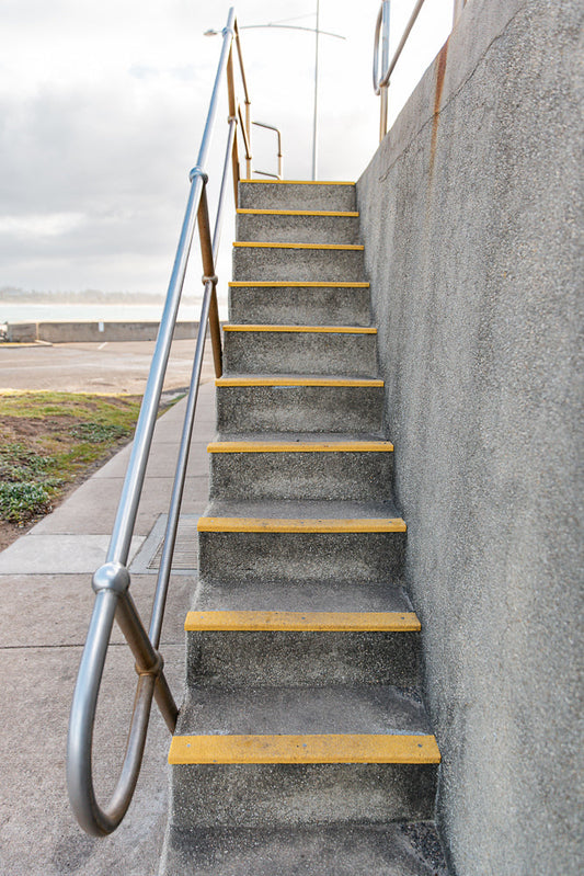 A concrete staircase with yellow anti-slip treads ascends alongside a textured concrete wall. A metal handrail with a curved section is visible on the left. The background shows a paved area, grass, and a body of water under a cloudy sky.