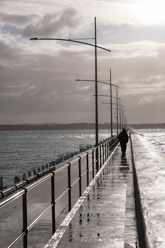 A lone figure walks along a wet pier under a cloudy sky. The pier is lined with railings and streetlights, and the water is choppy.