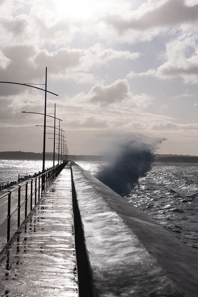 A wave crashes against the side of a pier on a cloudy day. The pier is wet and reflective, with a railing and lampposts visible. The ocean is choppy and dark.