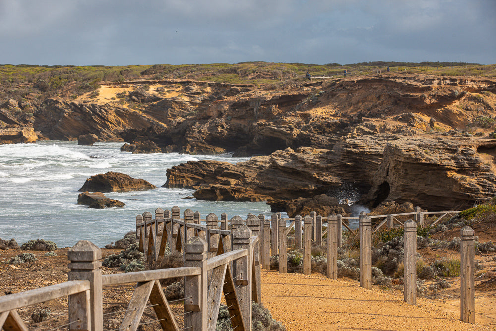 A wooden boardwalk winds along a rugged coastline with waves crashing against rocky cliffs under a cloudy sky. Low-lying coastal vegetation covers the hills.