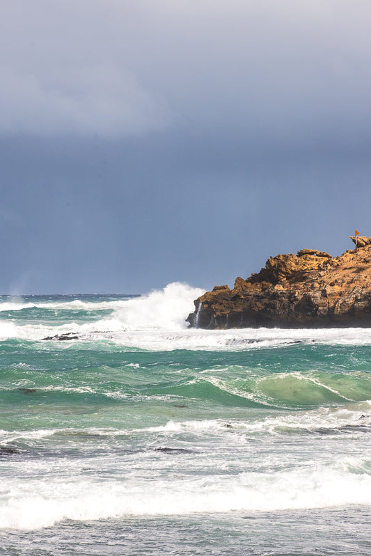 A dramatic seascape with large waves crashing against a rocky cliff face under a stormy sky. A yellow flag is visible on top of the cliff.