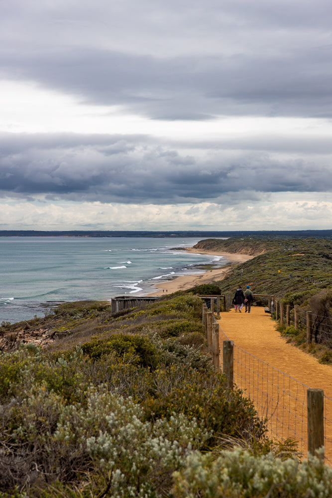 A couple walks along a sandy path beside a coastal reserve with the ocean and a cloudy sky in the background.