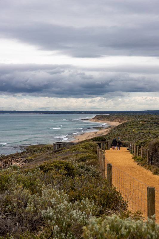 A couple walks along a sandy path beside a coastal reserve with the ocean and a cloudy sky in the background.