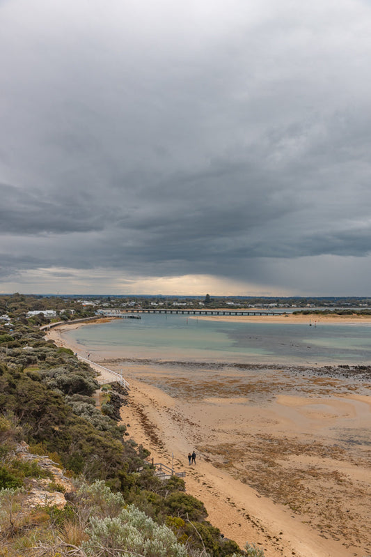 A wide shot of a coastal landscape under a dramatic, cloudy sky. A sandy beach curves along the water, with a boardwalk and lush vegetation bordering it. A long pier stretches across the calm, turquoise water towards a distant town.
