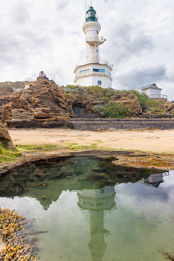 A white lighthouse with a green lantern room stands on a rocky outcrop overlooking the sea. A couple sits on the rocks in the foreground, and the lighthouse is reflected in a calm pool of water.