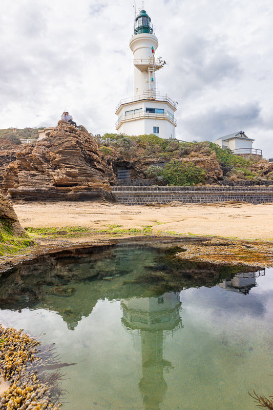 A white lighthouse with a green lantern room stands on a rocky outcrop overlooking the sea. A couple sits on the rocks in the foreground, and the lighthouse is reflected in a calm pool of water.