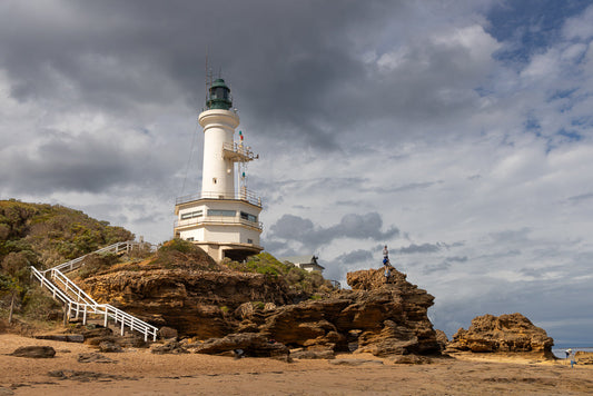 A white lighthouse with a green lantern room stands on a rocky outcrop overlooking a sandy beach. White stairs lead up the grassy hill to the lighthouse. The sky is filled with dramatic, dark clouds.