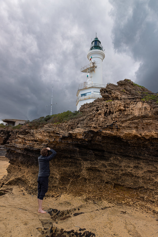 A person stands barefoot on a rocky shore, looking through binoculars at a lighthouse under a cloudy sky.