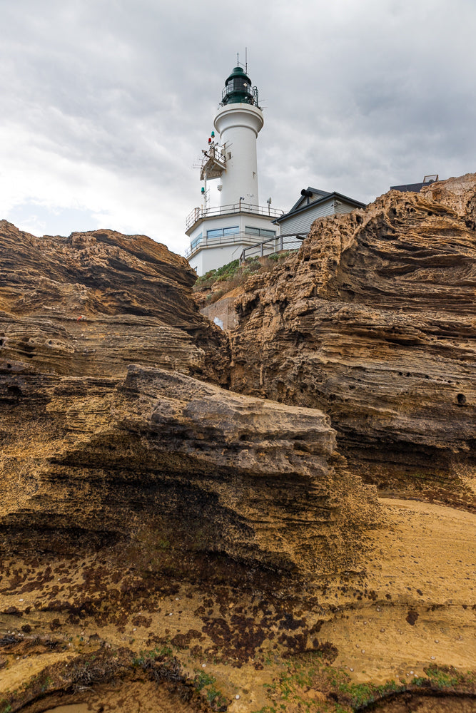 A white lighthouse with a green top stands tall against a cloudy sky, perched on a rocky cliffside. The foreground is dominated by textured, weathered rocks in shades of brown and yellow, with some patches of green vegetation visible.