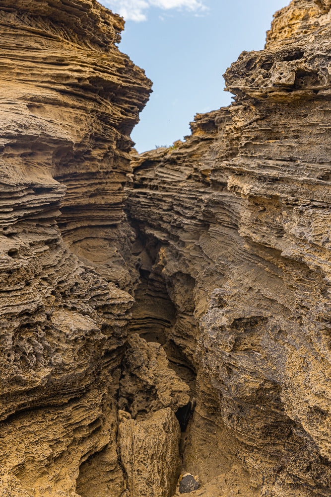 A narrow passage between weathered, layered rock formations. The textured, porous stone is a warm golden-brown color, with a sliver of blue sky visible at the top.