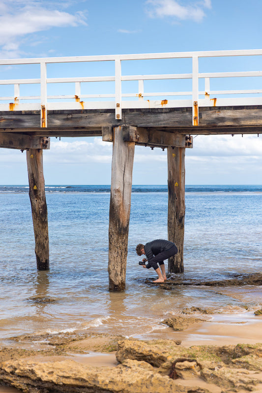 A person crouches on a rocky shore, taking a photo of a wooden pier support post in the water. The pier has a white railing and weathered wooden beams.