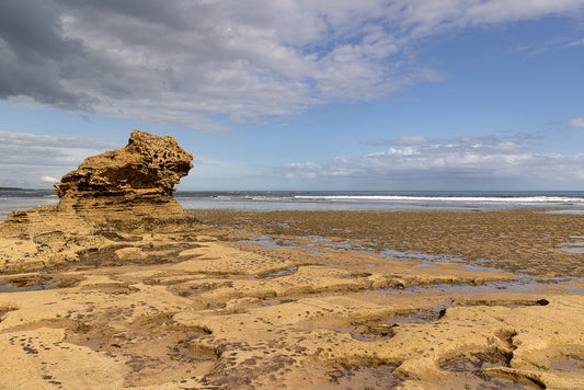 A large, weathered rock formation stands on a sandy beach at low tide. The textured sand is dotted with tide pools and seaweed. The ocean stretches to the horizon under a partly cloudy sky.