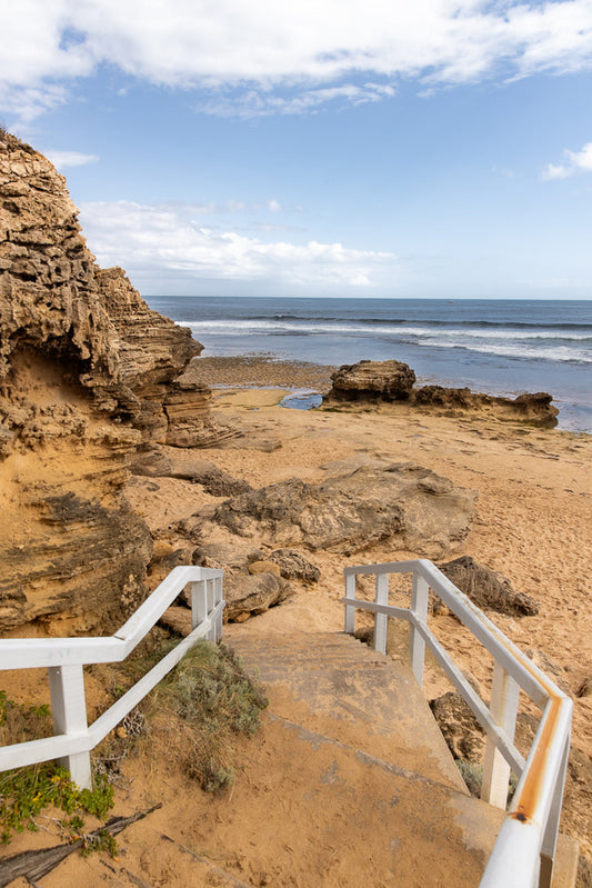 A set of weathered white stairs with rusty railings leads down to a sandy beach with rocky outcrops and the ocean under a partly cloudy sky. A small boat is visible on the horizon.