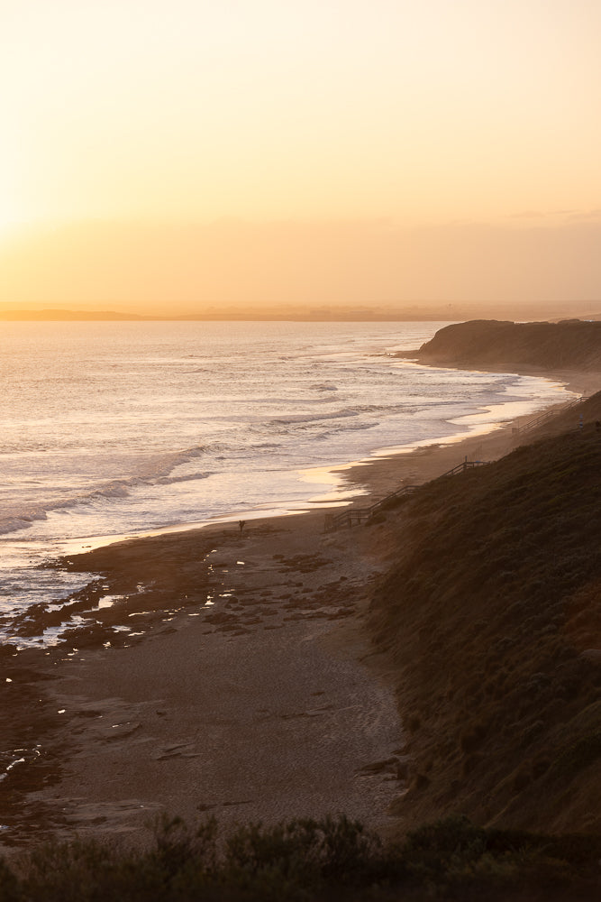 A wide shot of a beach at sunset. The sun is low on the horizon, casting a warm golden glow over the ocean and sky. Gentle waves roll onto the sandy shore, and a few people can be seen walking along the water's edge. A grassy cliff overlooks the beach, with a wooden staircase leading down to the sand.