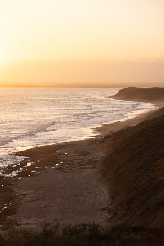 A wide shot of a beach at sunset. The sun is low on the horizon, casting a warm golden glow over the ocean and sky. Gentle waves roll onto the sandy shore, and a few people can be seen walking along the water's edge. A grassy cliff overlooks the beach, with a wooden staircase leading down to the sand.