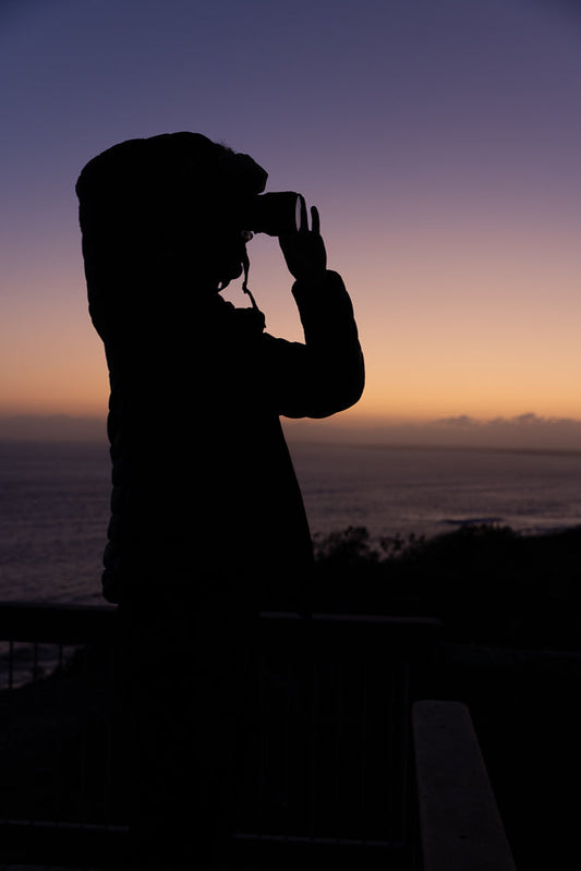 Silhouette of a person wearing a hooded jacket, looking through binoculars at the ocean during a purple and orange sunset.