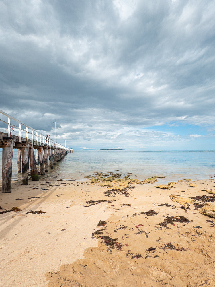 A long white pier extends into the calm ocean under a cloudy sky. The sandy beach in the foreground is scattered with rocks and seaweed.