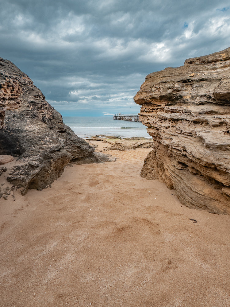 A view of a sandy beach with weathered rock formations in the foreground, leading to a calm ocean and a distant pier under a cloudy sky.