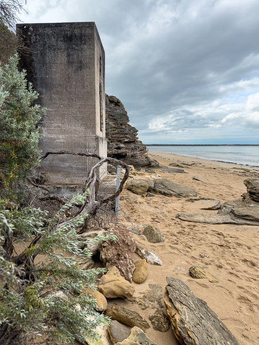A concrete structure stands on a sandy beach next to layered rock formations. A person is visible in the distance on the beach. The sky is overcast with clouds.