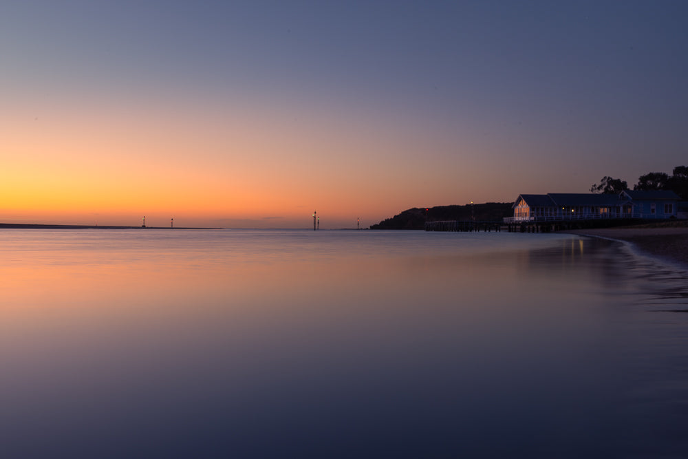 A serene twilight scene shows a calm body of water reflecting the gradient of the sky, transitioning from warm orange to deep blue. A long pier with a blue building on stilts extends into the water on the right, with lights illuminating the structure. Several navigation buoys are visible in the distance.
