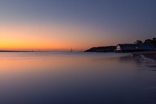 A serene twilight scene shows a calm body of water reflecting the gradient of the sky, transitioning from warm orange to deep blue. A long pier with a blue building on stilts extends into the water on the right, with lights illuminating the structure. Several navigation buoys are visible in the distance.