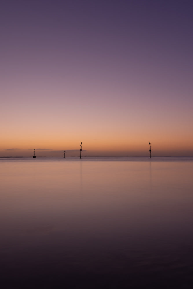 A serene seascape at twilight with a gradient sky transitioning from deep purple to soft orange. Several channel markers stand in the calm water, silhouetted against the horizon.