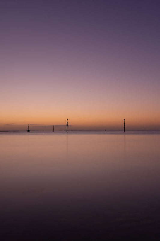 A serene seascape at twilight with a gradient sky transitioning from deep purple to soft orange. Several channel markers stand in the calm water, silhouetted against the horizon.