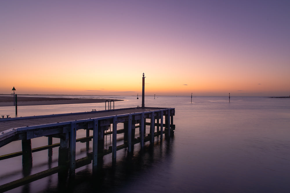 A wooden pier extends into calm water under a soft purple and orange sky at sunrise or sunset. The tranquil scene is enhanced by the gentle reflections on the water's surface and the distant navigational markers.