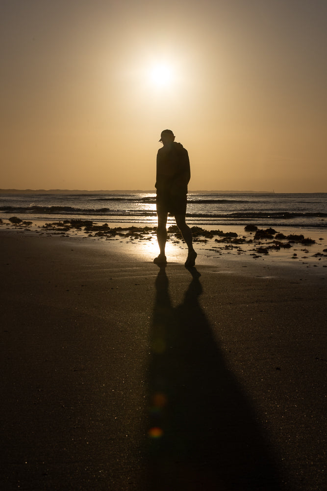 A person wearing a baseball cap and shorts walks along a sandy beach at sunset. The sun is low in the sky, casting a long shadow of the person onto the sand. Gentle waves lap at the shore in the background.