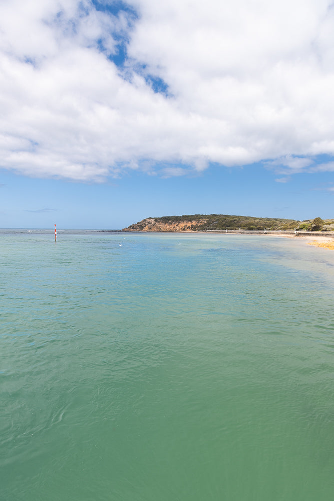 A calm, turquoise bay with a sandy beach and a tree-covered hill in the background under a partly cloudy sky. A red and white marker pole stands in the water near the shore.