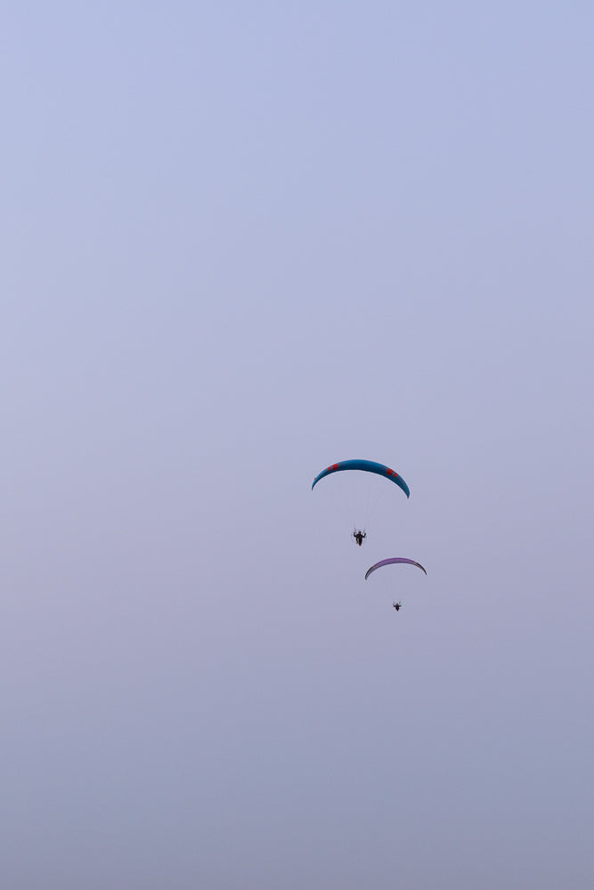 Two paragliders soar through a pale blue sky. The paraglider in front is blue with orange accents, and the one behind it is purple. Both paragliders are suspended in the air, appearing to be enjoying a peaceful flight.