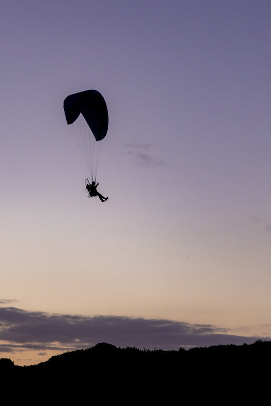 A silhouette of a paraglider in mid-air against a purple and orange twilight sky. The paraglider is suspended beneath a dark blue parachute, with the pilot visible in a harness. The dark silhouette of a hilly landscape with vegetation forms the bottom of the frame.