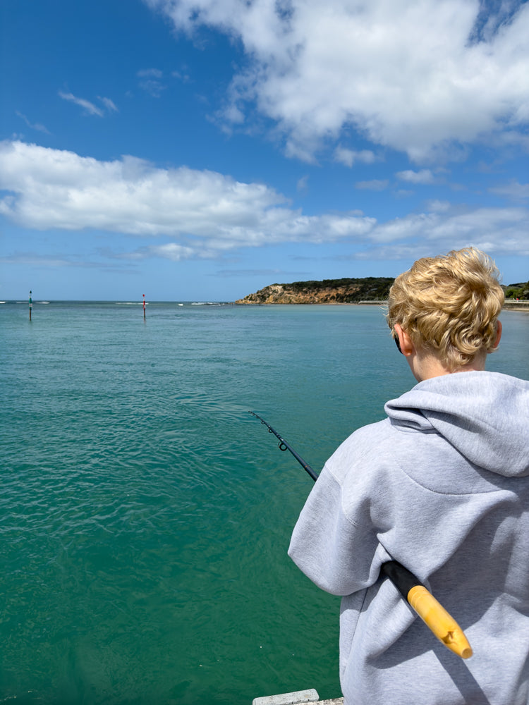 A young boy with blond hair stands on a wooden pier, facing away from the camera, holding a fishing rod. A tackle box and a plastic bag are on the pier beside him. The water is calm, and a long bridge is visible in the distance under a partly cloudy sky.