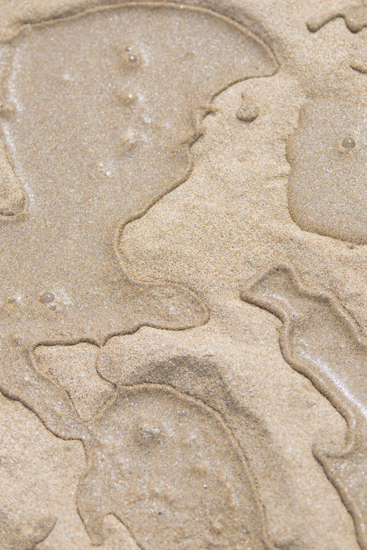 Close-up of wet sand with abstract patterns formed by receding water. The sand is light brown and textured, with shimmering highlights from the moisture. The patterns are organic and flowing, resembling natural contours or ripples.