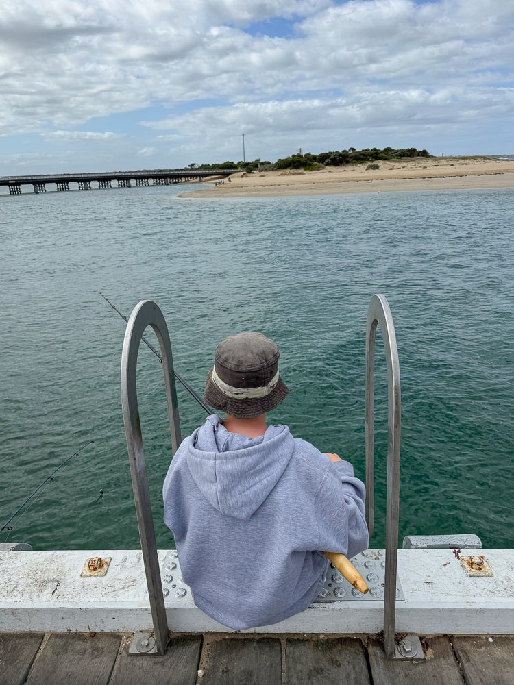 A person wearing a grey hoodie and a bucket hat sits on a pier, facing away from the camera, looking out at the water. A fishing rod is visible in the foreground.