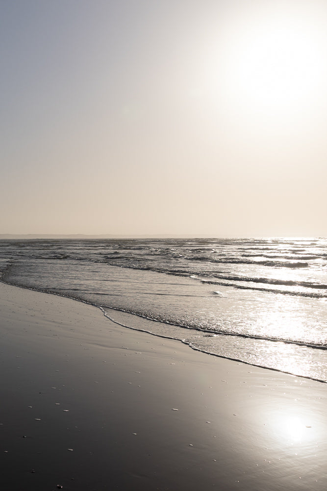 A serene beach scene with gentle waves lapping onto the shore under a hazy, sunlit sky. The wet sand reflects the soft light, creating a peaceful and tranquil atmosphere.