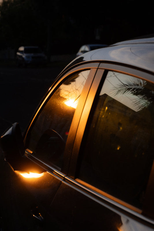 The setting sun casts a warm orange glow on the side of a dark car, reflecting in the window and on the side mirror. The scene is dark with the exception of the bright sunlight.