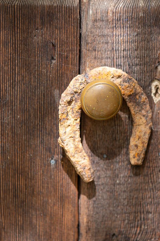 A rusty horseshoe is mounted on a weathered wooden door, serving as a door handle. The horseshoe is positioned horizontally, with its curved ends pointing upwards and downwards. A brass doorknob is attached to the center of the horseshoe.