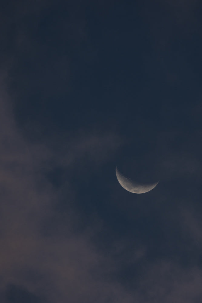 A crescent moon is visible in a dark, cloudy sky. The moon is illuminated from the right, showing its curved edge and textured surface.
