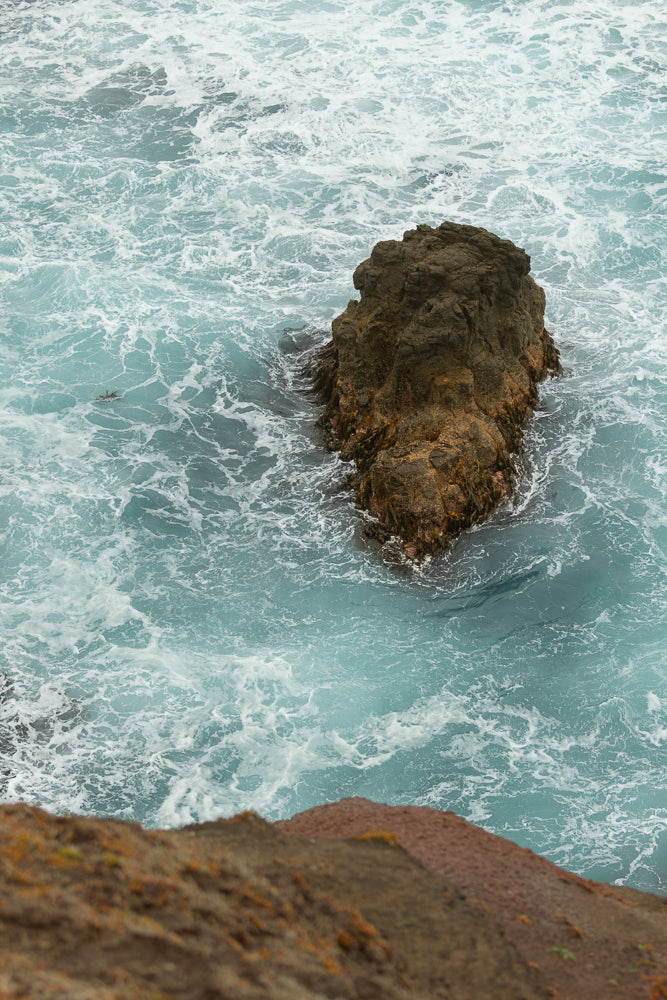 A large, dark rock formation stands in the middle of turquoise water with white foam. The water is choppy and turbulent, with waves crashing against the rock. The foreground shows a blurred view of a rocky cliff face.