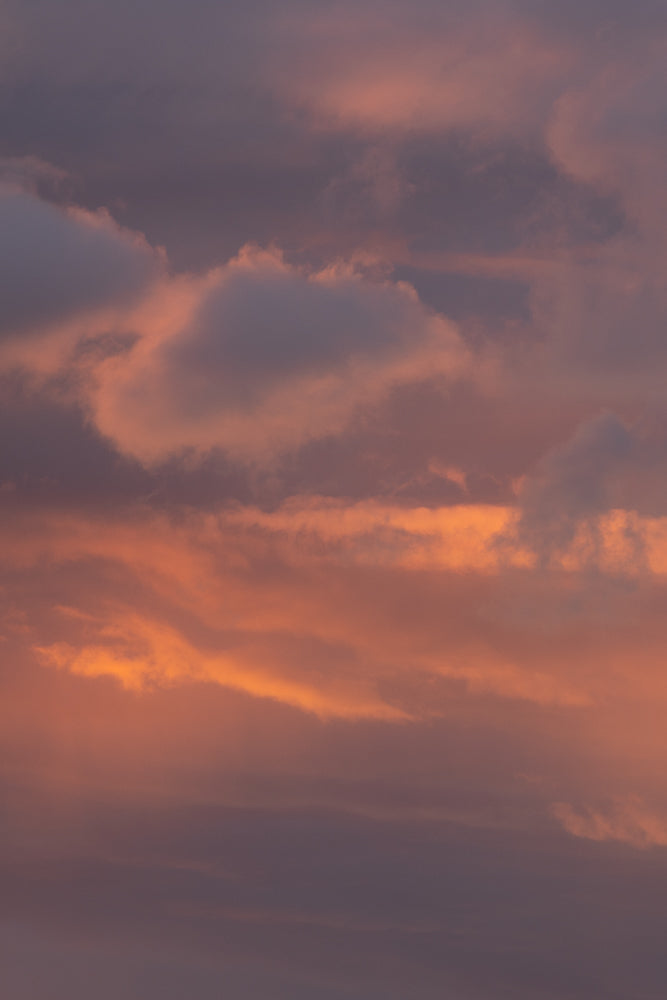 A sky filled with clouds illuminated by the warm glow of sunset. The clouds range in color from deep purple to soft pink and vibrant orange, creating a dramatic and beautiful natural display.