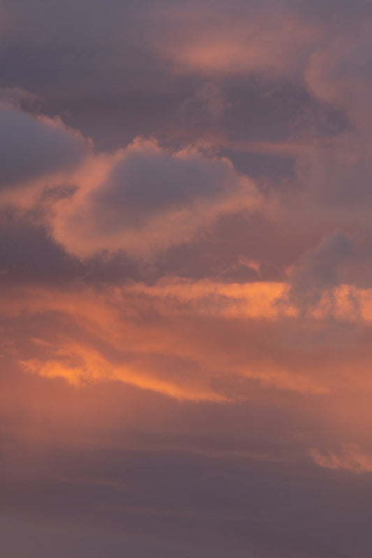 A sky filled with clouds illuminated by the warm glow of sunset. The clouds range in color from deep purple to soft pink and vibrant orange, creating a dramatic and beautiful natural display.