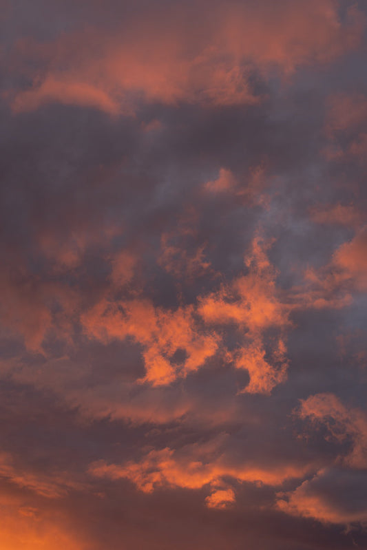 A dramatic sunset with fiery orange clouds interspersed with dark, moody clouds. The light catches the edges of the clouds, creating a vibrant and textured sky.