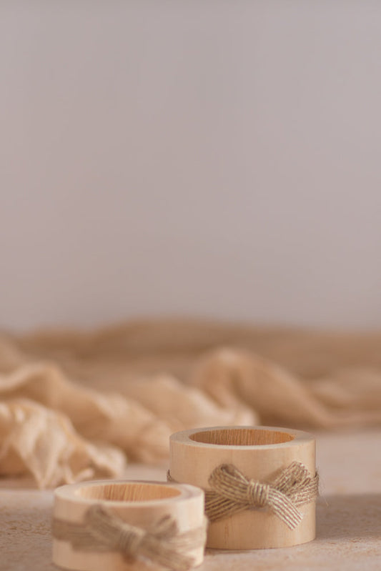 Two wooden napkin rings with twine bows sit on a textured surface, with a soft, draped fabric in the background.