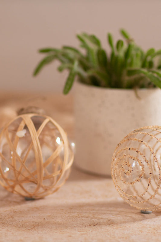 Two delicate glass Christmas ornaments sit on a textured surface. One is adorned with a spiral pattern of natural fibers, and the other is wrapped in thin, light-colored reeds.