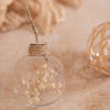 A close-up shot of a clear glass ornament filled with dried baby's breath flowers. The ornament is tied with twine and hangs against a soft, textured background in neutral tones.