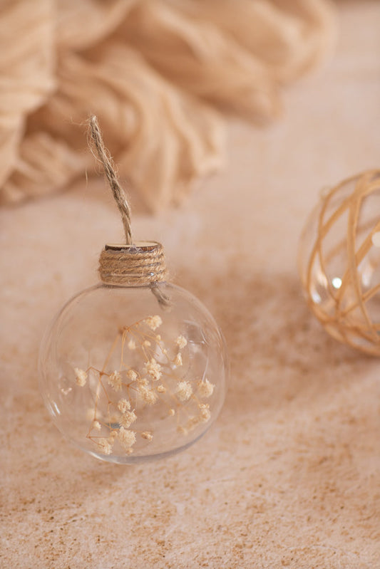 A close-up shot of a clear glass ornament filled with dried baby's breath flowers. The ornament is tied with twine and hangs against a soft, textured background in neutral tones.