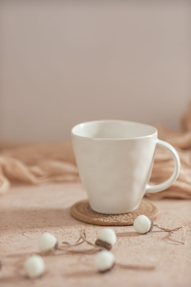 A white mug sits on a woven coaster on a textured surface. A string of small, white pom-pom acorns is draped in front of the mug.